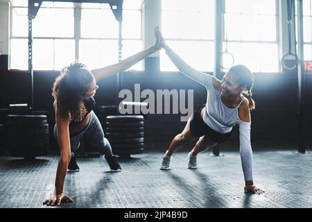 Motivation makes all the difference. two young women giving each other a high five while doing push ups at the gym. Stock Photo