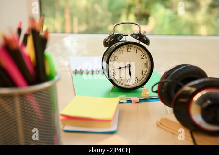 Focus on a black alarm clock on open notepad with school supplies and stationery on blurred foreground. Back to school Stock Photo