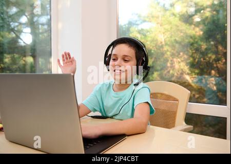 Schoolboy in headphones, waving with hand to the teacher while studying remotely from home, sitting at table with laptop Stock Photo