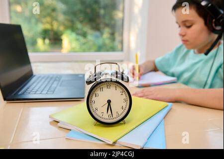 Focus on a black alarm clock on textbooks with studying schoolboy, watching online course on laptop on the background Stock Photo