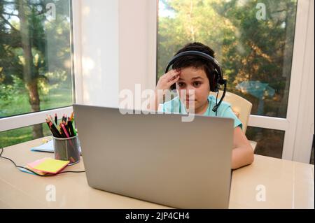 Adorable pupil of primary school in audio headset, watching online video lesson on laptop. Homeschooling. Online school Stock Photo