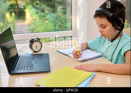 School boy having online classes, sitting at home in quarantine, using laptop and taking notes, watching online lessons Stock Photo