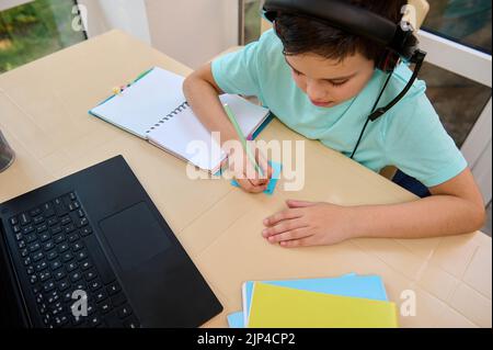 Schoolboy having online classes, at home in quarantine, using laptop and taking notes, watching online lessons. Top view Stock Photo