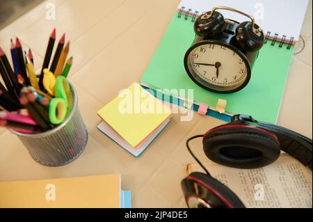 Top view black alarm clock on open notepad with school supplies and stationery on blurred foreground. Back to school Stock Photo