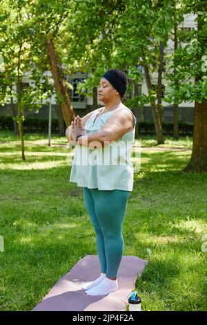 Vertical full length portrait of overweight black woman doing yoga outdoors in green park and meditating with eyes closed Stock Photo