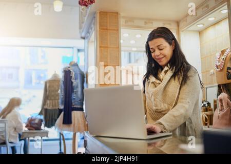 Using online resources to manage her business. an attractive mature female entrepreneur working on a laptop in her self-owned boutique. Stock Photo