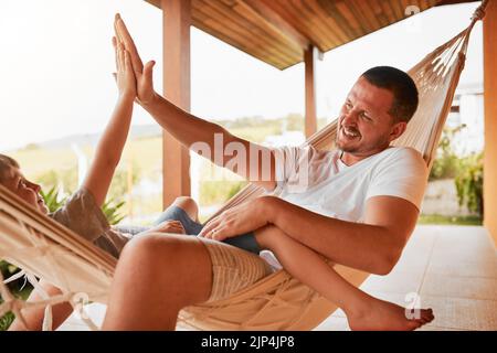 Hes more than just a father. an mature man and his young son high fiving while lying outside on a hammock. Stock Photo