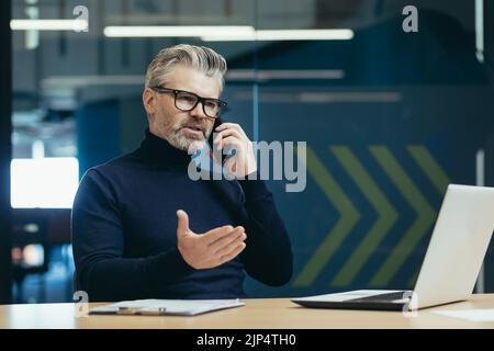 Senior and mature boss businessman working inside office building, man thoughtful and serious talking on the phone, gray haired investor in glasses working using laptop Stock Photo
