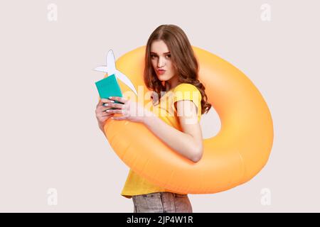 Portrait of tourist teenager girl in yellow T-shirt holding rubber ring, passport document and airplane mockup, looking at camera with pouts lips. Indoor studio shot isolated on gray background. Stock Photo