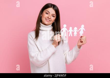 Smiling positive girl holds white paper family chain, looking at camera with happy facial expression, wearing white casual style sweater. Indoor studio shot isolated on pink background. Stock Photo