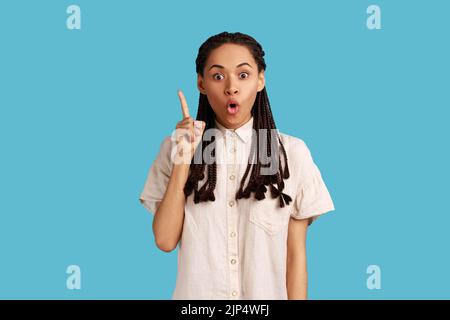 Pleased young woman with black dreadlocks pointing upwards, looking at camera with open mouth, having new great idea, wearing white shirt. Indoor studio shot isolated on blue background. Stock Photo