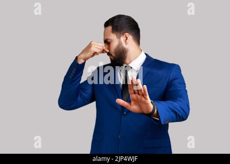 Bearded man grabbing nose, grimacing in disgust and showing stop gesture, expressing repulsion to stink, fart gases, wearing official style suit. Indoor studio shot isolated on gray background. Stock Photo