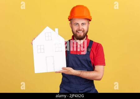 Portrait of worker man holding paper house, offering help in relocation, home repair services and maintenance, wearing overalls and cap. Indoor studio shot isolated on yellow background. Stock Photo