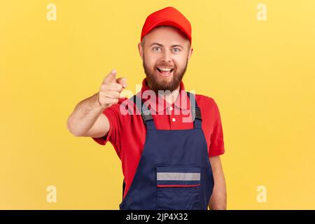 Portrait of smiling bearded satisfied man worker standing and pointing finger to camera, choosing you, wearing overalls and red cap. Indoor studio shot isolated on yellow background. Stock Photo