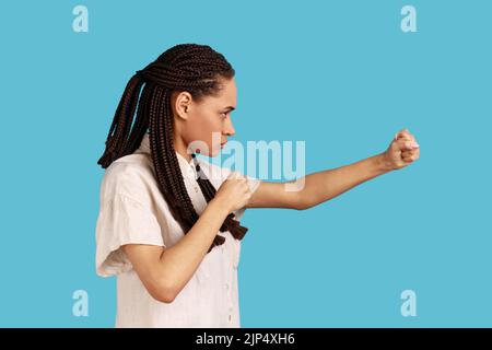 Side view portrait of woman with black dreadlocks having serious look, keeping fists clenched, defencing herself from offence and bad treatment. Indoor studio shot isolated on blue background. Stock Photo