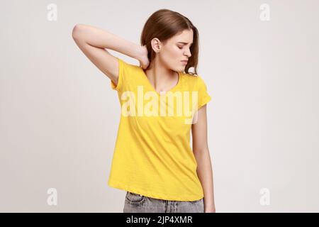 Shoulders and neck pain. Portrait of tired upset teen girl in yellow casual style T-shirt standing massaging neck to relieve pain, muscle strain in back. Indoor studio shot isolated on gray background Stock Photo