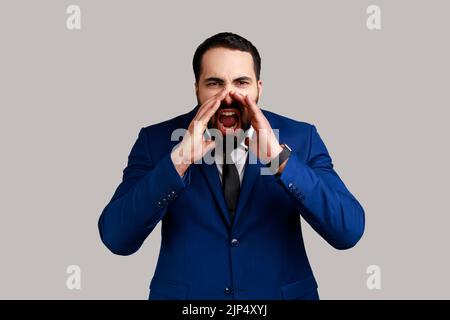 Attention. Portrait of stressed furious bearded man standing, holding arms near wide open mouth and screaming, wearing official style suit. Indoor studio shot isolated on gray background. Stock Photo