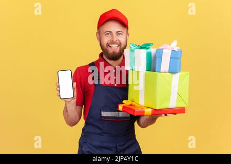 Portrait of smiling delivery man wearing blue overalls holding stack of present boxes and smart phone with blank screen for advertisement. Indoor studio shot isolated on yellow background. Stock Photo