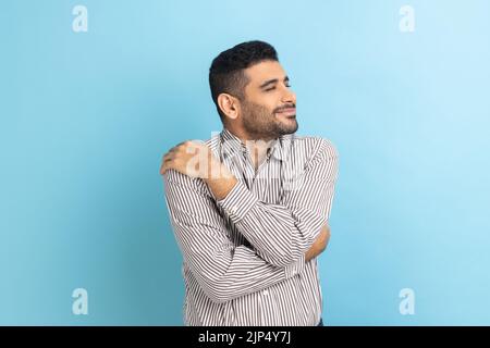 Portrait of happy handsome businessman with beard standing and hugging himself with toothy smile enjoying, positive self-esteem, wearing striped shirt. Indoor studio shot isolated on blue background. Stock Photo