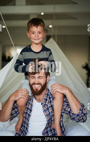 Riding high on dads shoulders. Cropped portrait of an adorable little boy sitting on his dads shoulders while sitting on the bed at home. Stock Photo