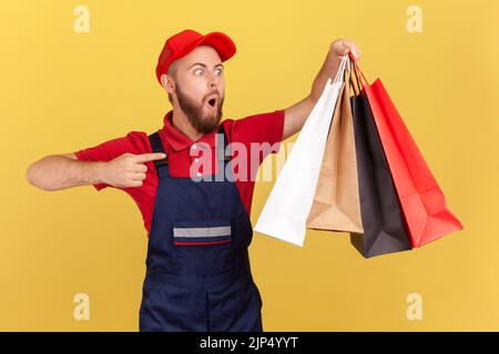 Portrait of surprised shocked delivery man in uniform pointing finger at shopping bags in his hand, fast delivery from mall, service industry. Indoor studio shot isolated on yellow background. Stock Photo