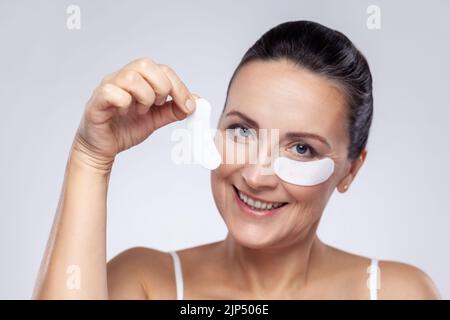 Beauty treatment concept. Closeup portrait of middle aged woman applies patches under eyes, removes wrinkles and dark circles, has manicure. Indoor studio shot isolated on gray background. Stock Photo