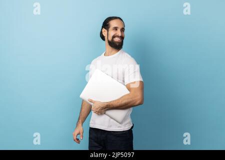 Side view of positive man with beard wearing white T-shirt standing, holding closed laptop or folder, looking back with optimistic expression. Indoor studio shot isolated on blue background. Stock Photo