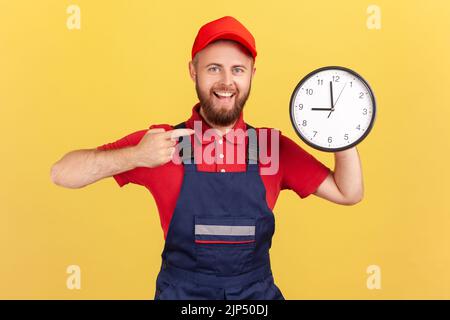 Portrait of delighted man worker standing and pointing at big wall clock in his hands, looking at camera, wearing overalls and red cap. Indoor studio shot isolated on yellow background. Stock Photo