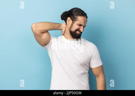 Portrait of man with beard wearing white T-shirt touching neck, feeling acute pain, suffering spine problems, osteochondrosis, frowning face.Indoor studio shot isolated on blue background. Stock Photo