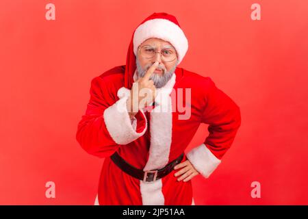 Elderly man with gray beard wearing santa claus costume touching nose, gesturing you are liar, being distrustful of talk, suspecting falsehood. Indoor studio shot isolated on red background. Stock Photo