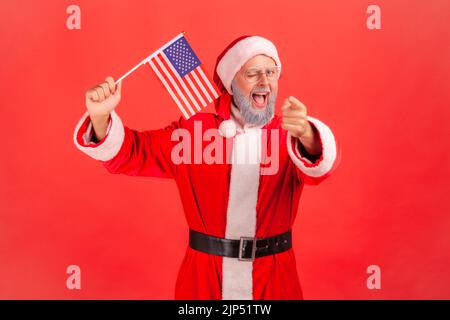 Satisfied elderly man with gray beard wearing santa claus costume waving USA flag winking and pointing to camera, keeps mouth open. Indoor studio shot isolated on red background. Stock Photo