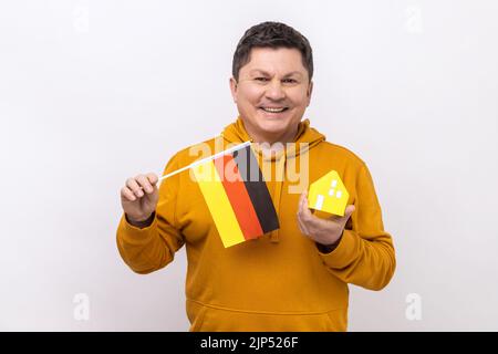 Optimistic man holding German flag and paper house, dreaming to buy his own apartrment in Germany, looking at camera, wearing urban style hoodie. Indoor studio shot isolated on white background. Stock Photo