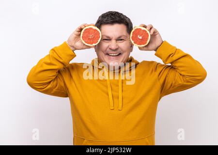 Portrait of smiling middle aged man having fun, holding two halves of grapefruit, expressing positive emotions, wearing urban style hoodie. Indoor studio shot isolated on white background. Stock Photo