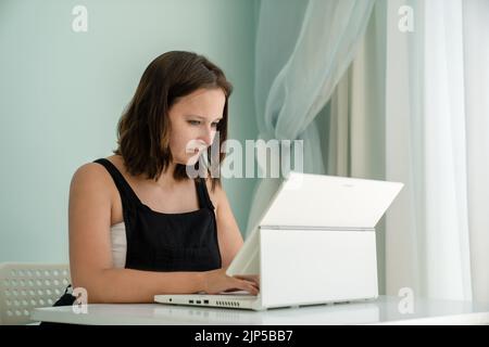 Girl is sitting in front of her laptop transformer at desk in her house. Concept of remote work and education, side view Stock Photo