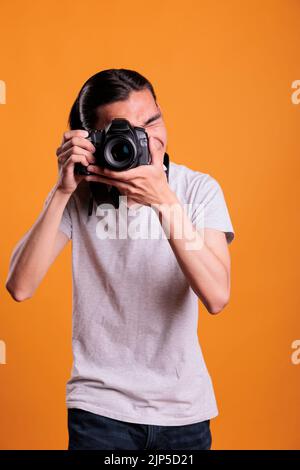 Photographer taking photos on professional digital camera, looking in viewfinder. Young asian man using photographing equipment, front view medium shot, creative hobby, photoshoot Stock Photo