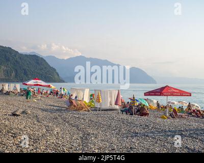 Gonio, Georgia. 08.03.2022  Pebble public beach on the Black Sea coast. Lots of people on the water. People on deck chairs and under umbrellas. Hot ev Stock Photo