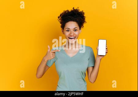 Amazed shocked african american curly young woman, surprised looks at the camera, holds cell phone with a blank white screen and points finger at it, stand on isolated orange background. Mock-up Stock Photo