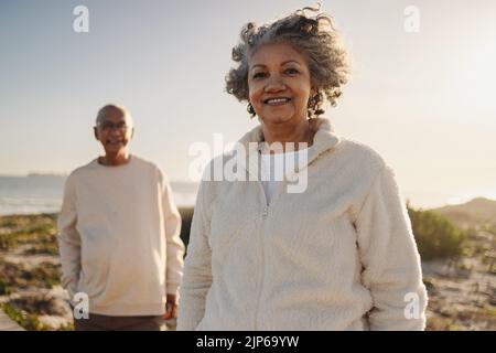Elderly woman smiling at the camera while standing on a foot bridge at the beach. Happy senior woman enjoying a refreshing holiday with her husband af Stock Photo