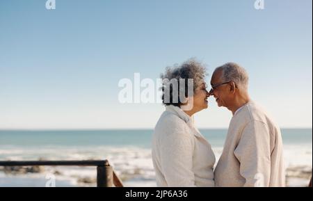 Being romantic by the ocean. Happy senior couple smiling and touching their noses together while standing on a seaside foot bridge. Retired elderly co Stock Photo