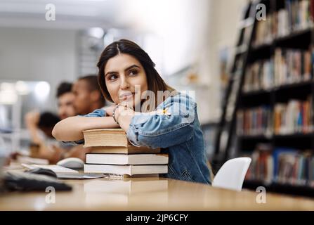 Shes got the whole world in her sights. a young woman resting on a pile of books in a college library and looking thoughtful. Stock Photo