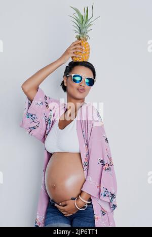 My baby will love pineapples as much as I do. Studio shot of a beautiful young pregnant woman holding a pineapple on top of her head against a gray Stock Photo