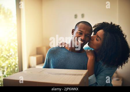Look at my strong man. Portrait of a cheerful young man carrying a cardboard box while his girlfriend gives him a kiss on the cheek. Stock Photo