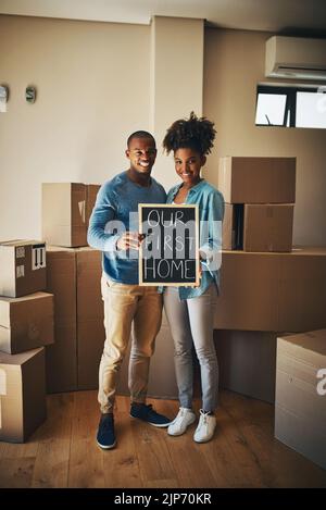 This is where our life begins. Portrait of a cheerful young couple holding up a sign saying our first home together while being surrounded by Stock Photo