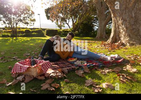 Happy caucasian couple lying on rug having picnic in sunny autumn garden and drinking wine Stock Photo