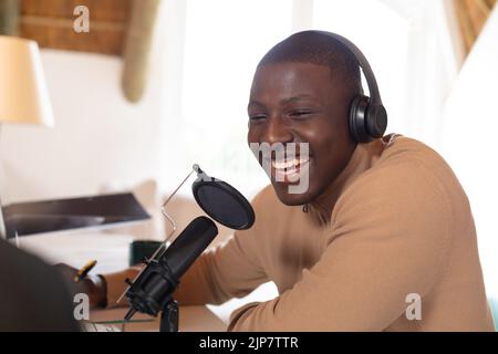 Laughing african american man in headphones making podcast at home using computer and microphone Stock Photo