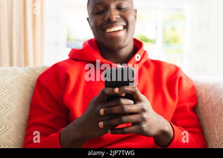 Laughing african american man sitting on couch at home using on smartphone Stock Photo