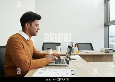 Concentrated young Indian entrepreneur working on laptop in modern office, checking reports and e-mails Stock Photo