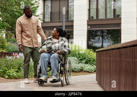 Full length portrait of happy black couple with young woman in wheelchair enjoying walk in city garden together and looking at each other with love, c Stock Photo
