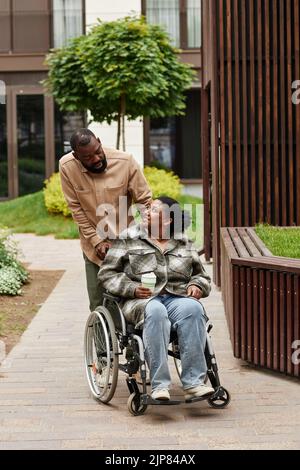 Vertical full length portrait of black couple with young woman in wheelchair enjoying walk in city garden and looking at each other Stock Photo