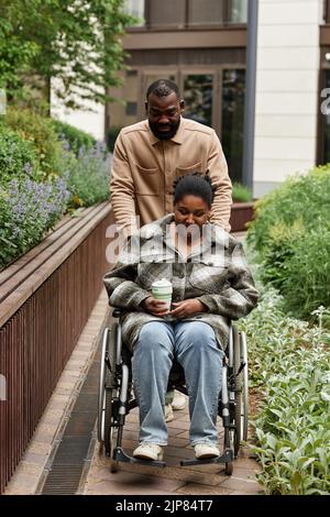 Vertical full length portrait of black young woman in wheelchair outdoors in city garden with smiling husband assisting Stock Photo
